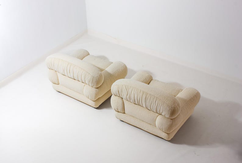 May include: Two cream-coloured, upholstered ottomans. Each ottoman has a rectangular base with rounded corners and a series of rounded, cushioned sections. The fabric has a textured appearance, and the ottomans are placed on a white surface.