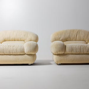 May include: Two cream-coloured, upholstered armchairs. Each chair has a rounded back and armrests, with a pleated design on the back and seat. The chairs sit on small, silver-coloured feet, against a white background.