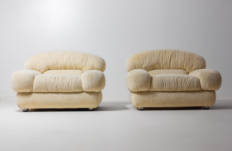 May include: Two cream-coloured, upholstered armchairs. Each chair has a rounded back and armrests, with a pleated design on the back and seat. The chairs sit on small, silver-coloured feet, against a white background.