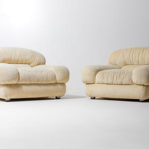May include: Two cream-coloured upholstered armchairs. Each chair features a rounded back and armrests, with a pleated texture. The chairs sit on low, square bases with small, silver feet. The background is plain white.