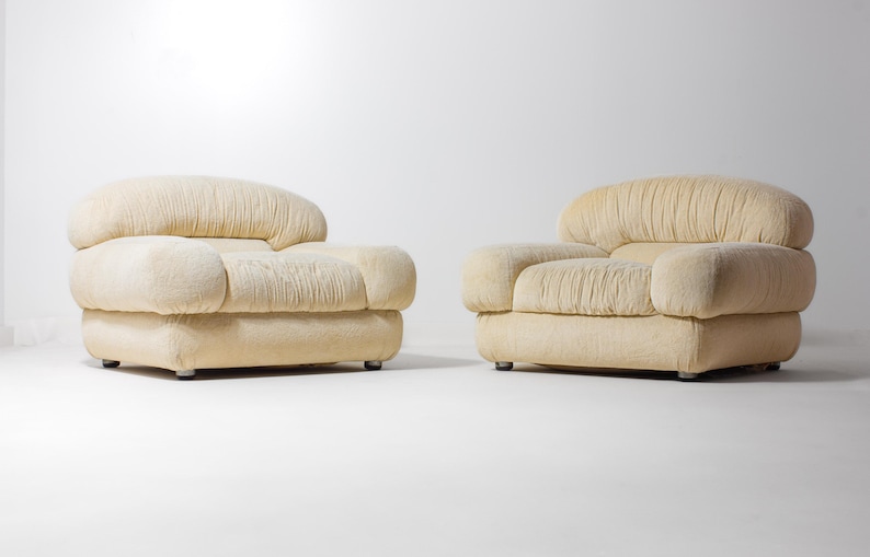 May include: Two cream-coloured upholstered armchairs. Each chair features a rounded back and armrests, with a pleated texture. The chairs sit on low, square bases with small, silver feet. The background is plain white.