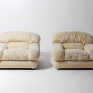 May include: A pair of cream-coloured armchairs. Each chair features a rounded back and armrests, with a pleated texture on the back and seat. The chairs are set against a white background.
