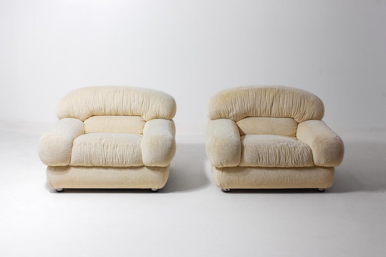 May include: A pair of cream-coloured armchairs. Each chair features a rounded back and armrests, with a pleated texture on the back and seat. The chairs are set against a white background.
