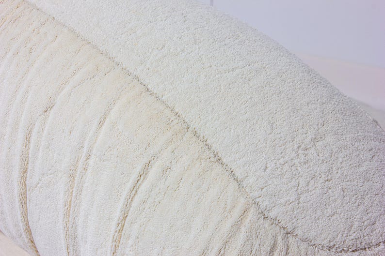 May include: Close-up of a textured, off-white cushion. The cushion has a soft, plush appearance with a series of curved, stitched lines running along its surface. The fabric has a slightly uneven texture, adding to its cozy look.