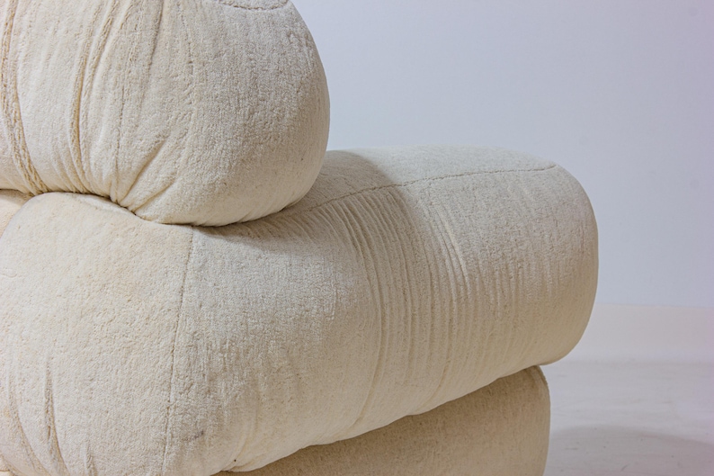 May include: Close-up of a cream-coloured, modular sofa. The sofa has a textured, fabric-like appearance with rounded, cylindrical shapes. The design features visible stitching and a soft, inviting aesthetic, suitable for a living room.