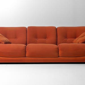 May include: A three-seat orange sofa with rounded armrests and two matching throw pillows. The sofa has a low profile and a tufted backrest. The cushions are a solid orange color, and the overall design is modern.