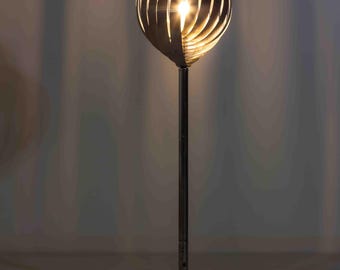 May include: A modern floor lamp with a unique design. The lamp features a silver metal base and a tall, slender pole supporting a light fixture. The light fixture is an oval shape with curved, slotted metal pieces, creating a warm glow.
