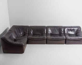 Może przedstawiać: Ciemnobrązowa skórzana sofa modułowa z narożnikiem i trzema pojedynczymi siedziskami. Sofa ma niski profil i lekko zużyty wygląd, sugerujący styl vintage. Skóra ma błyszczący połysk.