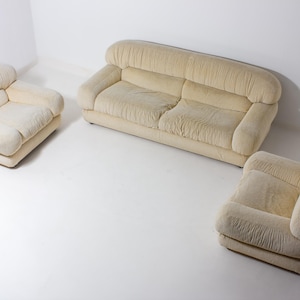 May include: A three-piece beige sofa suite. The set comprises a two-seater sofa and two armchairs. The furniture features a rounded design with soft, textured upholstery. The pieces are arranged on a white surface, creating a clean and minimalist aesthetic.