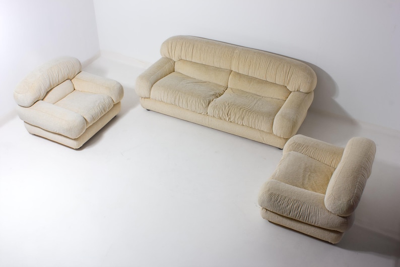 May include: A three-piece beige sofa suite. The set comprises a two-seater sofa and two armchairs. The furniture features a rounded design with soft, textured upholstery. The pieces are arranged on a white surface, creating a clean and minimalist aesthetic.