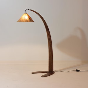 Puede incluir: Una lámpara de pie de madera con un diseño curvo y arqueado. La lámpara tiene una pantalla triangular con una superficie ámbar texturizada. La base es de color marrón oscuro y la lámpara está enchufada a una toma de corriente. Altura aproximada 180 cm.
