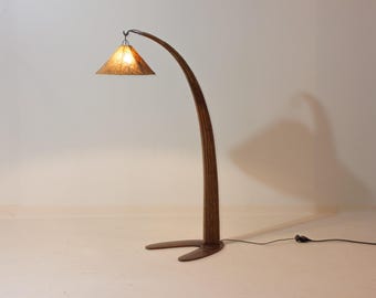 May include: A wooden floor lamp with a curved, arching design. The lamp has a triangular lampshade with a textured, amber surface. The base is dark brown, and the lamp is connected to a power outlet. The lamp's height is approximately 180 cm.