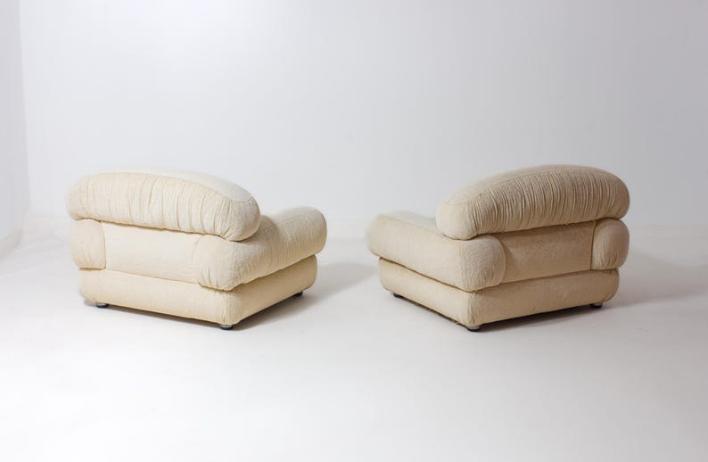 May include: Two cream-coloured armchairs with rounded backs and armrests. The backs have a pleated design, and the chairs sit on a white surface. The chairs are a neutral colour and would fit in many home decor styles.