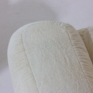 May include: Close-up of a section of a cream-coloured upholstered armchair. The image highlights the rounded armrest and the textured fabric. The stitching along the edges is visible, adding detail to the design. The neutral colour suggests a versatile piece of furniture.