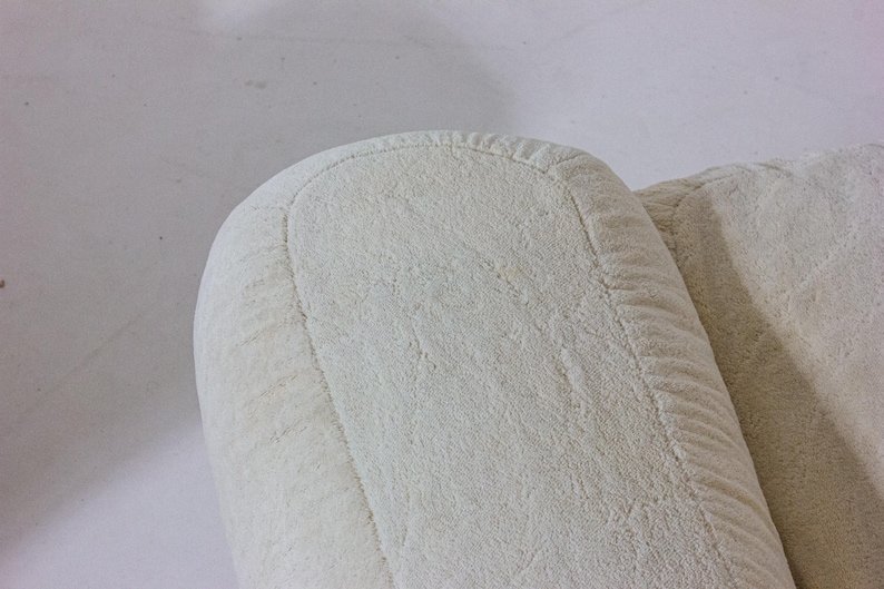 May include: Close-up of a section of a cream-coloured upholstered armchair. The image highlights the rounded armrest and the textured fabric. The stitching along the edges is visible, adding detail to the design. The neutral colour suggests a versatile piece of furniture.