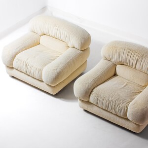 May include: Two cream-coloured armchairs. Each chair has a rounded back and armrests, with a square seat. The fabric has a textured appearance. The chairs are placed on a white surface.