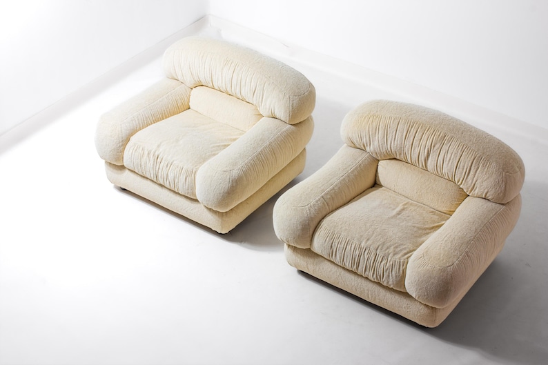 May include: Two cream-coloured armchairs. Each chair has a rounded back and armrests, with a square seat. The fabric has a textured appearance. The chairs are placed on a white surface.