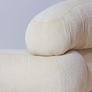 May include: Close-up of a section of a cream-coloured sofa. The sofa has a rounded back and armrest, with a textured fabric. The design is modern and minimalist, suitable for home decor.