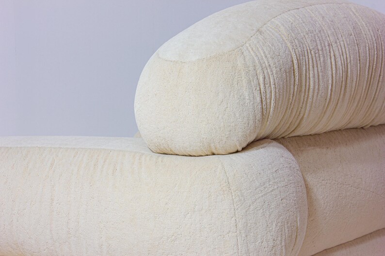 May include: Close-up of a section of a cream-coloured sofa. The sofa has a rounded back and armrest, with a textured fabric. The design is modern and minimalist, suitable for home decor.