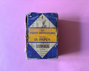 Può includere: Mazzo di carte dei tarocchi vintage intitolato "Le Tarot Divinatoire" del Dr. Papus. La scatola è blu e gialla con testo e illustrazioni in francese. Il mazzo appare invecchiato con usura, su sfondo rosa.
