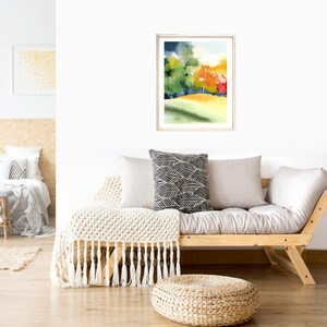 May include: A watercolor painting of a forest scene with green, yellow, orange, and red trees. The painting is framed in a light wood frame and is hanging on a white wall above a light wood futon.