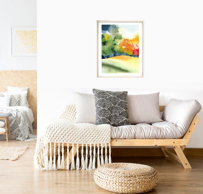 May include: A watercolor painting of a forest scene with green, yellow, orange, and red trees. The painting is framed in a light wood frame and is hanging on a white wall above a light wood futon.