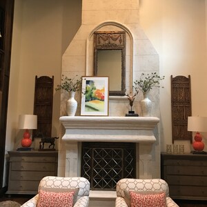 May include: A living room with a fireplace, two armchairs with patterned fabric and red pillows, and two lamps with red shades. The fireplace has a white stone surround and a black metal grate. There are two decorative wooden screens on either side of the fireplace.