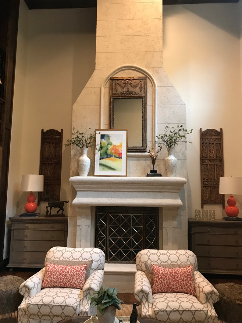 May include: A living room with a fireplace, two armchairs with patterned fabric and red pillows, and two lamps with red shades. The fireplace has a white stone surround and a black metal grate. There are two decorative wooden screens on either side of the fireplace.