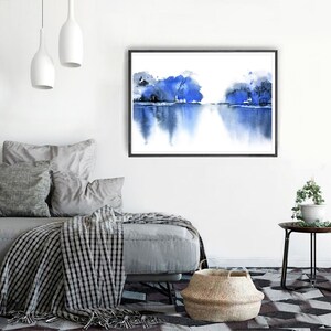 Op de afbeelding: Een aquarel schilderij van een blauw landschap met bomen die in het water worden weerspiegeld. Het schilderij is ingelijst in een zwart frame en hangt aan een witte muur.