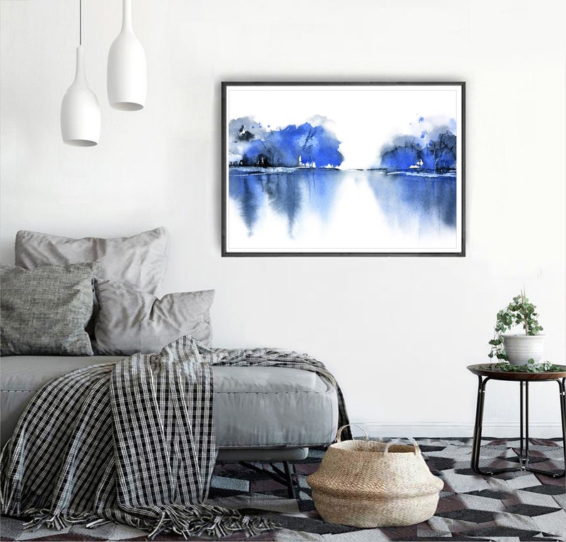 Op de afbeelding: Een aquarel schilderij van een blauw landschap met bomen die in het water worden weerspiegeld. Het schilderij is ingelijst in een zwart frame en hangt aan een witte muur.