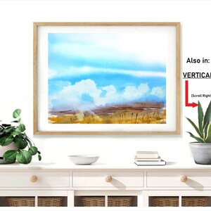 May include: A framed watercolor painting of a landscape with a blue sky, white clouds, and a field of tall grass. The painting is in a light wood frame and is hanging on a white wall.