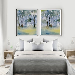 May include: Two framed watercolor paintings of a forest scene with blue and green hues. The paintings are hanging on a white wall above a gray bed with white pillows.