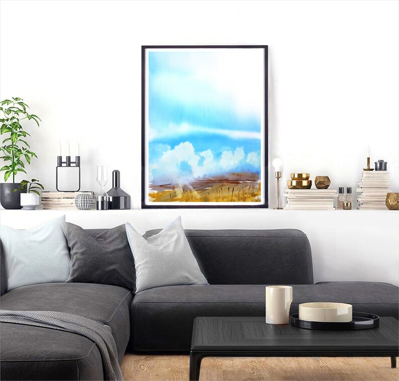 May include: A watercolor painting of a landscape with a blue sky, white clouds, and a field of golden grass. The painting is framed in a black frame and is hanging on a white wall.