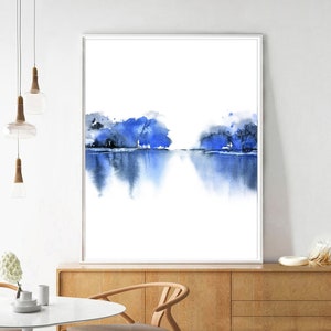 Op de afbeelding: Abstracte aquarelverf van een blauw landschap met bomen die in het water worden weerspiegeld. Het schilderij is ingelijst en hangt aan een witte muur.