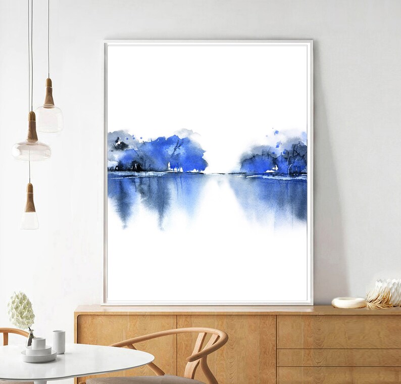 Op de afbeelding: Abstracte aquarelverf van een blauw landschap met bomen die in het water worden weerspiegeld. Het schilderij is ingelijst en hangt aan een witte muur.