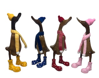 Könnte beinhalten: Vier hölzerne Entenfiguren, die Winteraccessoires tragen. Jede Ente hat einen Hut und einen Schal in einer anderen Farbe, und sie tragen alle gelbe, rote, blaue oder rosa Stiefel.