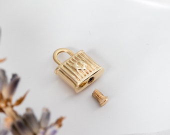 May include: A small, gold-colored padlock with a screw lying next to it. The padlock has a rectangular body with vertical lines and a small loop. A decorative element is visible on the front of the padlock.