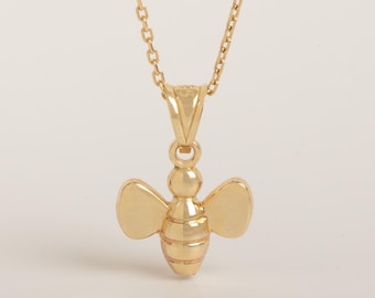 May include: A gold-colored necklace featuring a pendant shaped like a bee. The pendant has a segmented body and two smooth, rounded wings on each side. It hangs from a delicate, thin gold-colored chain with small, uniform circular links.