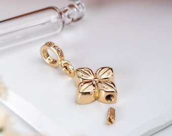 May include: A gold-colored, four-petal flower pendant with a small screw-on component. The pendant has a loop with small clear stones. A clear glass vial is in the background. The pendant is on a white surface.
