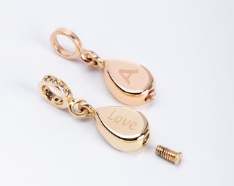 May include: Two gold teardrop-shaped charms with a screw closure. The charms are engraved with the letters "A" and "Love". The charms are shown with a variety of other charms, including a cross, key, wing, anchor, bone, and hammer. The charms are available in 14k and 18k gold.