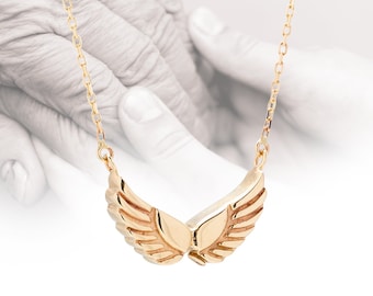 May include: A gold-colored necklace with a pendant shaped like stylized wings. The wings are detailed with layered lines, and the chain is delicate. The background features a blurred image of hands.