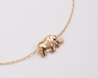 May include: A gold-colored necklace featuring a small, detailed elephant pendant. The elephant is facing left, with its trunk curled upwards. The chain is delicate and appears to be made of the same gold material. The background is plain white.
