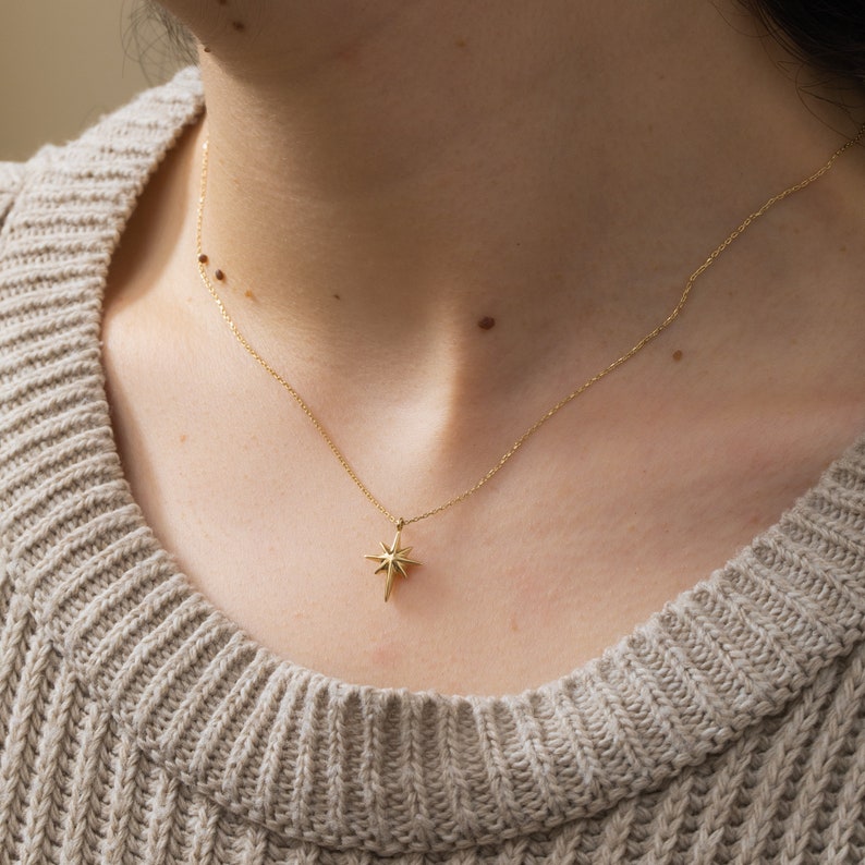 May include: A gold chain necklace with a star-shaped pendant. The necklace is worn by a person with a light brown sweater.