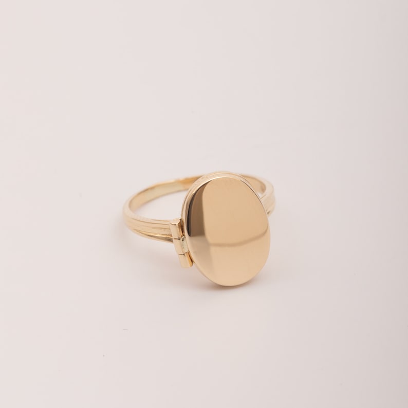 May include: A gold oval locket ring with a hinged opening. The ring is made of polished gold and has a smooth, shiny finish.