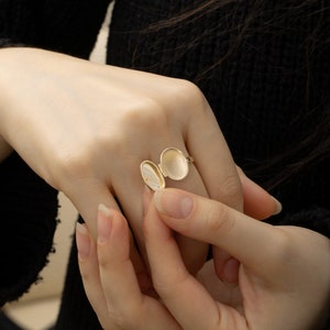 May include: A gold oval locket ring with the lid open. The ring is on a person's finger.