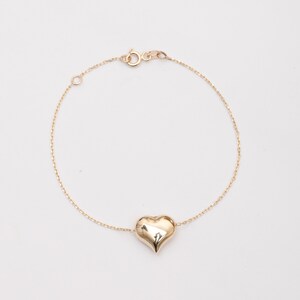 Puede incluir: Un brazalete de cadena de oro con un dije en forma de corazón. El corazón es liso y brillante.