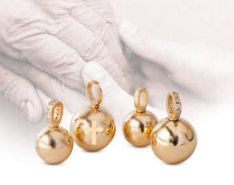 May include: Four gold-colored pendants with a spherical design. One pendant features a cross, and two have a loop adorned with small, clear stones. The pendants are displayed against a blurred background of hands.