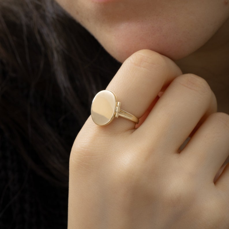 May include: A gold oval ring with a hinged top. The ring is on a person's finger.
