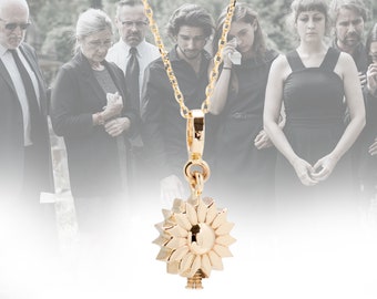 May include: A gold necklace with a sunflower pendant. The pendant has a keyhole in the center. The necklace is hanging against a blurred background of people in black clothing.