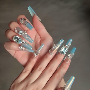Könnte beinhalten: Eine Hand, die lange, Sarg-förmige Acrylnägel zeigt. Die Nägel sind in einem Farbverlauf aus Hellblau und Klar lackiert, verziert mit silbernen Strasssteinen und großen, funkelnden Edelsteinen. Das Design ist aufwendig und glamourös.